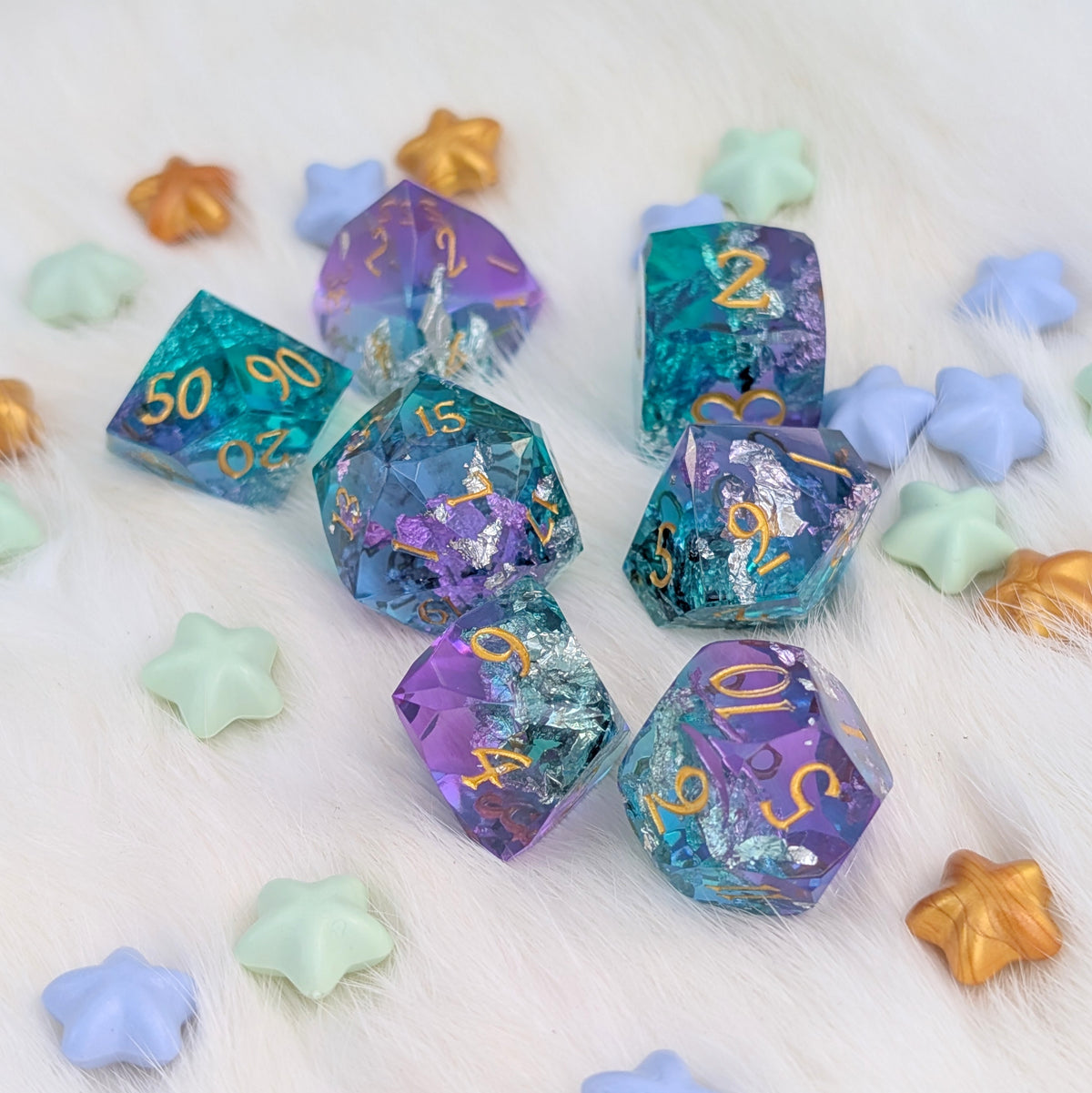 A set of translucent polyhedral gaming dice in teal, purple, and blue with gold numbers and foil accents, arranged on a white faux-fur surface with pastel star-shaped props.