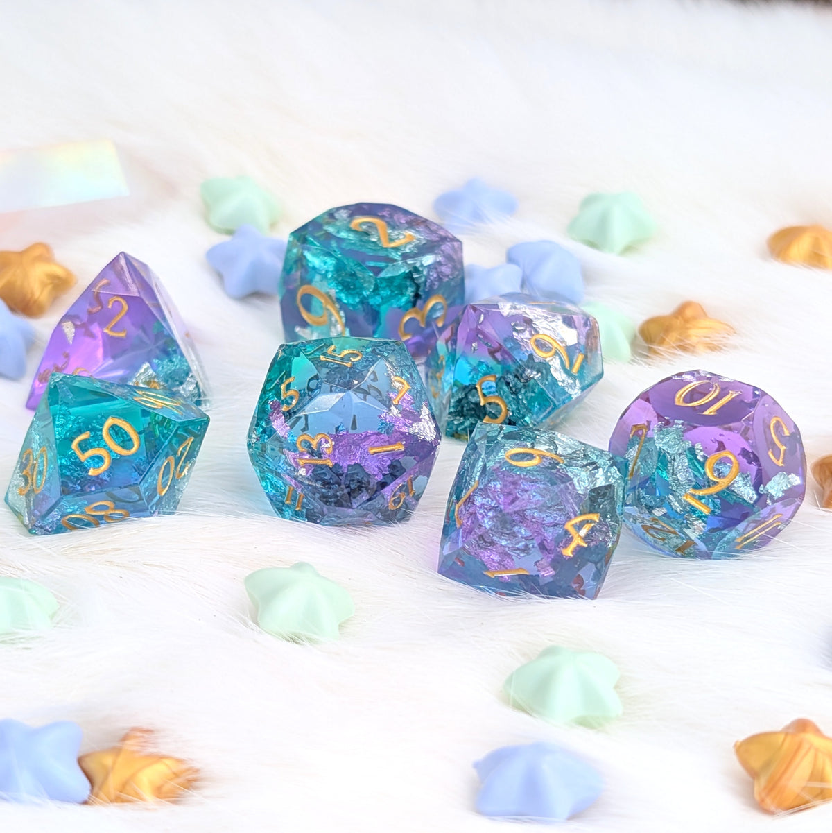 A set of holographic polyhedral gaming dice in pastel rainbow colors with gold numbering, arranged on a white faux fur surface with scattered pastel star-shaped confetti.