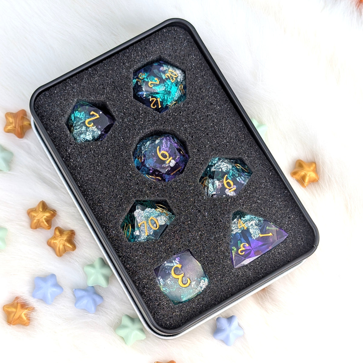 A set of seven iridescent d20 polyhedral dice with gold lettering, displayed in a black foam tray inside a black metal tin. The dice feature colorful, shifting glitter patterns of teal, purple, and blue.