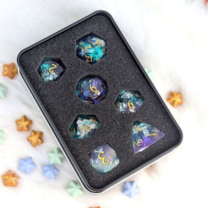 A set of seven iridescent d20 polyhedral dice with gold lettering, displayed in a black foam tray inside a black metal tin. The dice feature colorful, shifting glitter patterns of teal, purple, and blue.