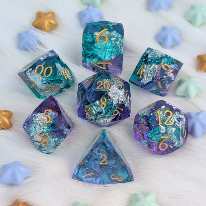 A set of seven polyhedral gaming dice with a glittery, iridescent finish in teal, purple, and blue tones, featuring gold-colored numbering, arranged on a white faux fur surface with pastel star-shaped props.