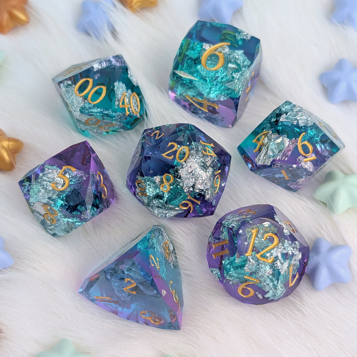 A set of seven iridescent polyhedral gaming dice with gold numbers, arranged on a white faux fur background with pastel star-shaped props.