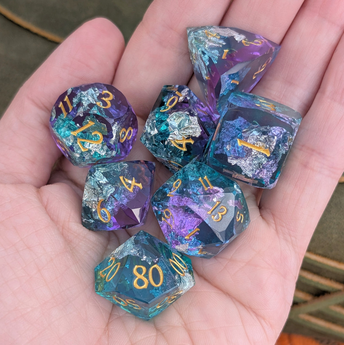 A set of seven iridescent polyhedral gaming dice resting in a person's hand. The dice feature a shimmering teal, purple, and blue coloration with metallic gold numbering.