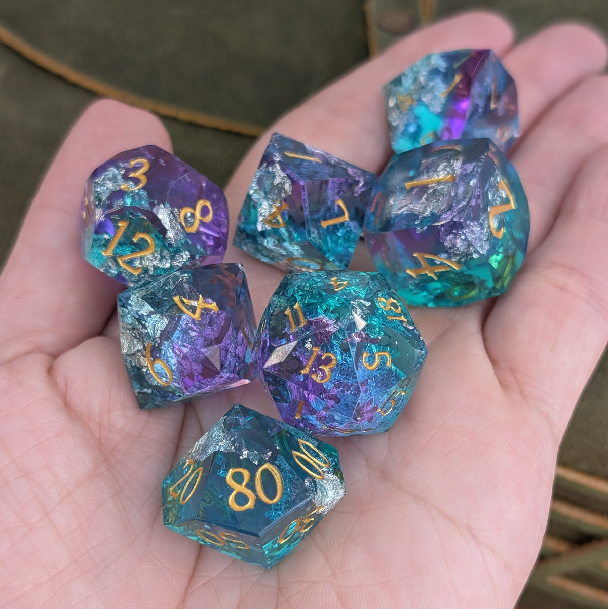 A set of translucent polyhedral gaming dice with gold numbers, displayed in a person's hand. The dice feature a dual-color iridescent finish shifting between purple and teal-blue, with some showing hints of green and pink.