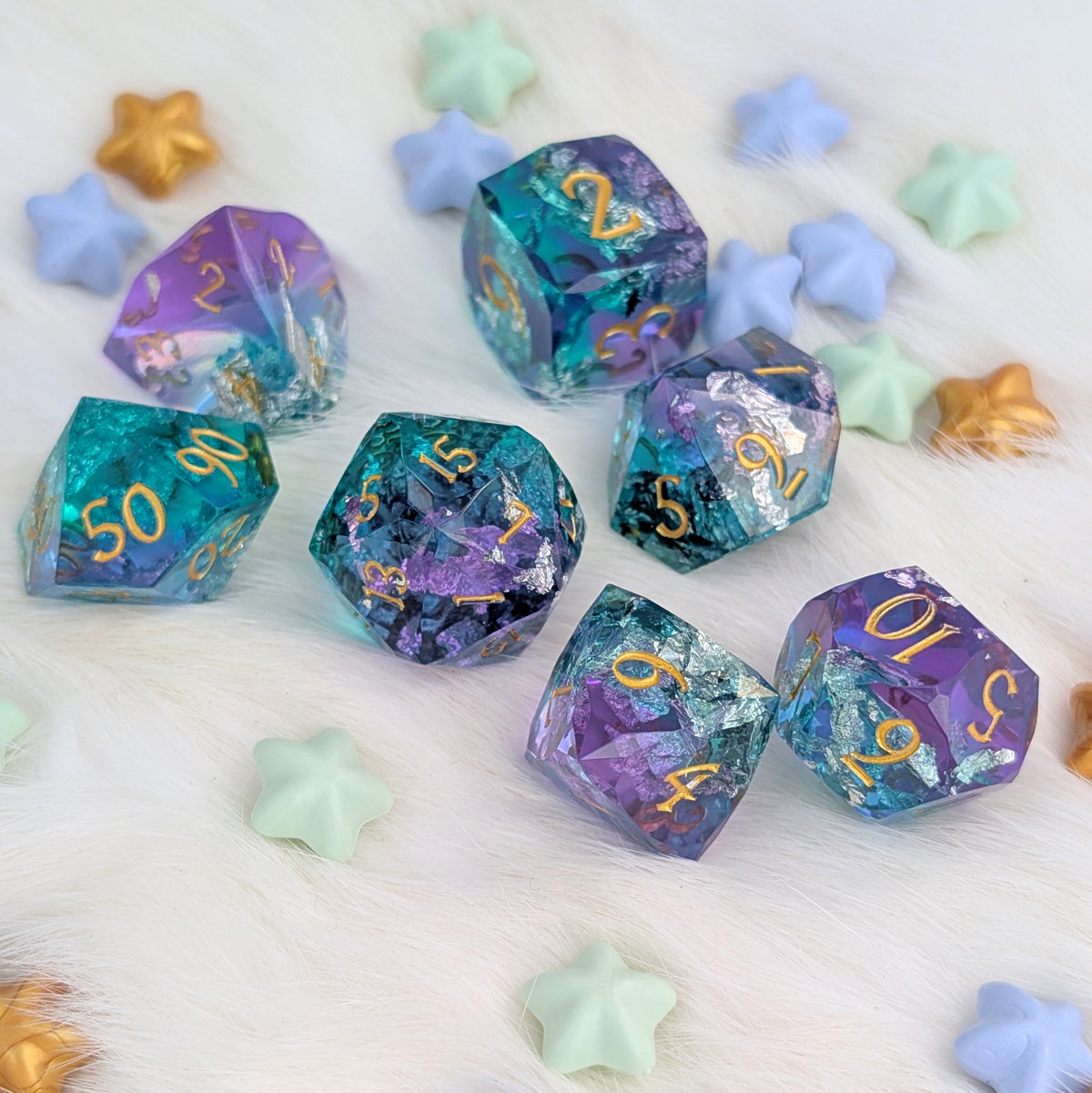 A set of translucent polyhedral gaming dice with gold numbers, featuring a purple, teal, and blue glitter pattern, arranged on a white faux fur surface with pastel star-shaped props.
