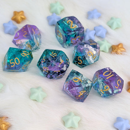 A set of translucent polyhedral gaming dice with gold numbers, featuring a purple, teal, and blue glitter pattern, arranged on a white faux fur surface with pastel star-shaped props.