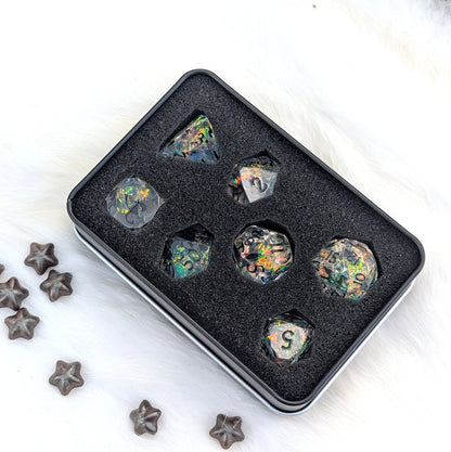 A set of seven iridescent resin polyhedral gaming dice displayed in a black foam-lined metal tin, with several dark star-shaped beads placed on a white faux-fur surface.