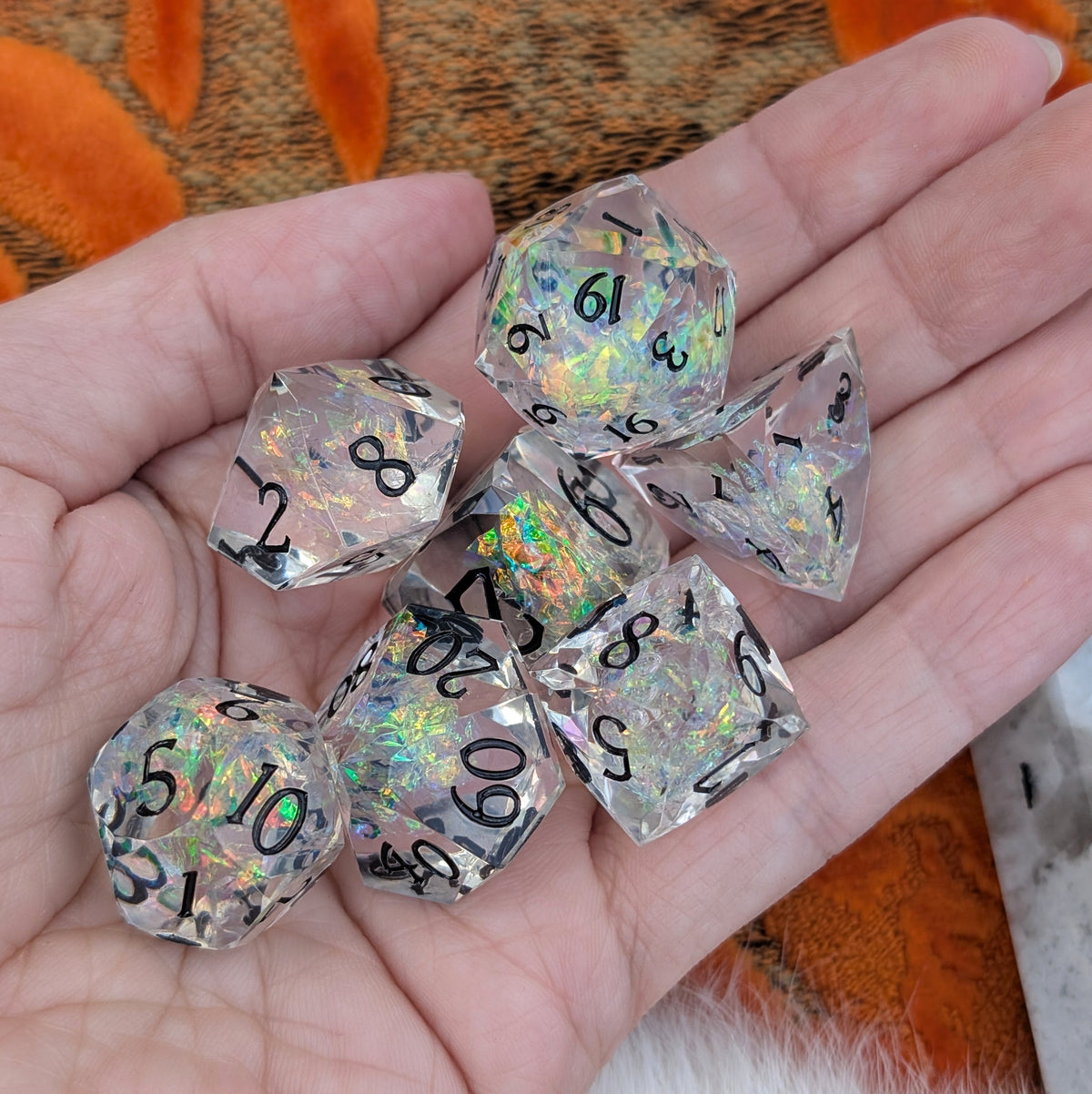 A set of clear holographic polyhedral dice resting in a person's hand. The dice feature black printed numbers and shimmer with iridescent rainbow reflections.