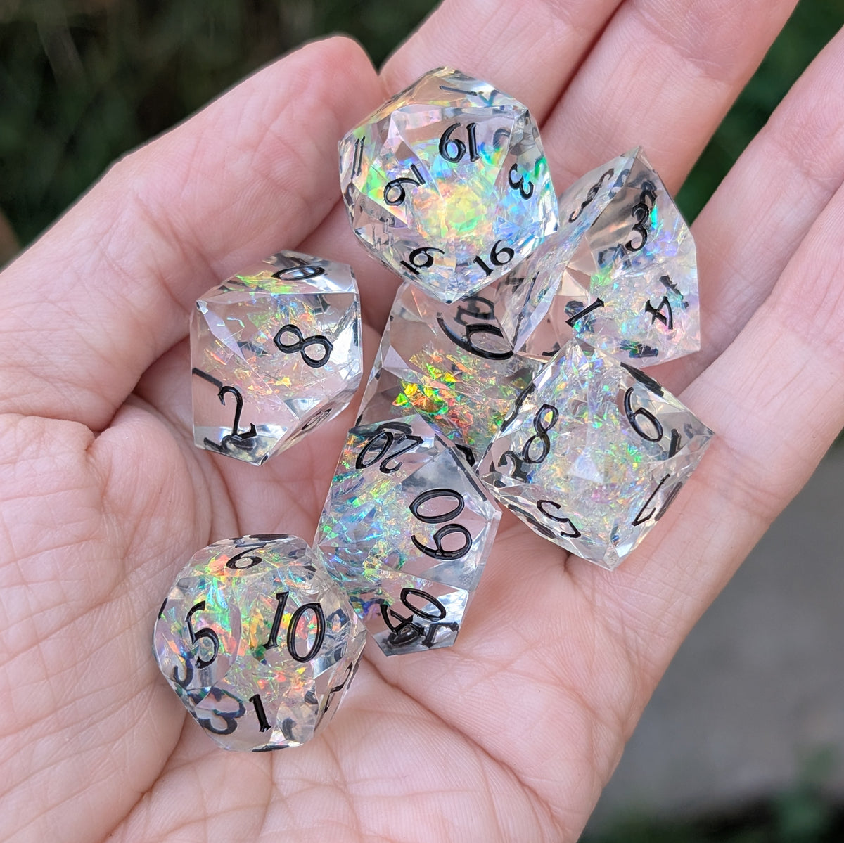 A set of clear acrylic polyhedral dice with black printed numbers and an iridescent holographic finish, held in a person's hand outdoors.