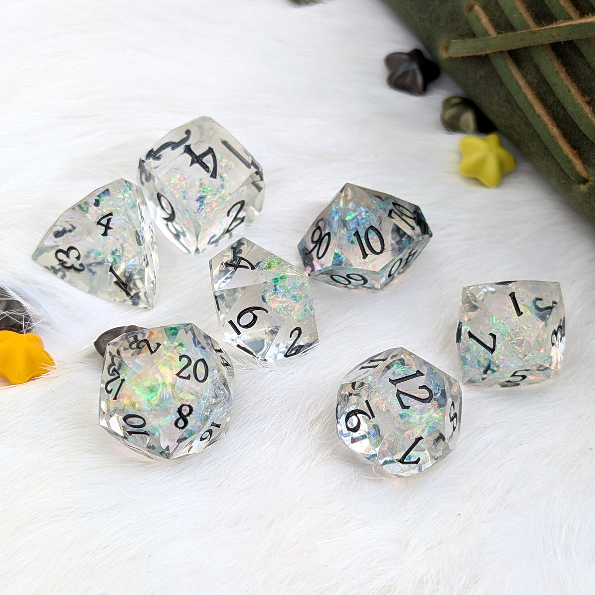 A set of clear acrylic polyhedral dice with black printed numbers and iridescent glitter, arranged on a white faux-fur surface with small decorative props.