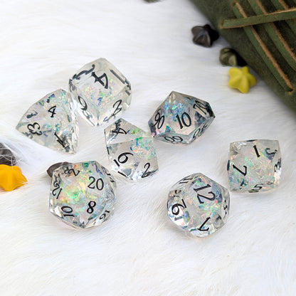 A set of clear acrylic polyhedral dice with black printed numbers and iridescent glitter, arranged on a white faux-fur surface with small decorative props.