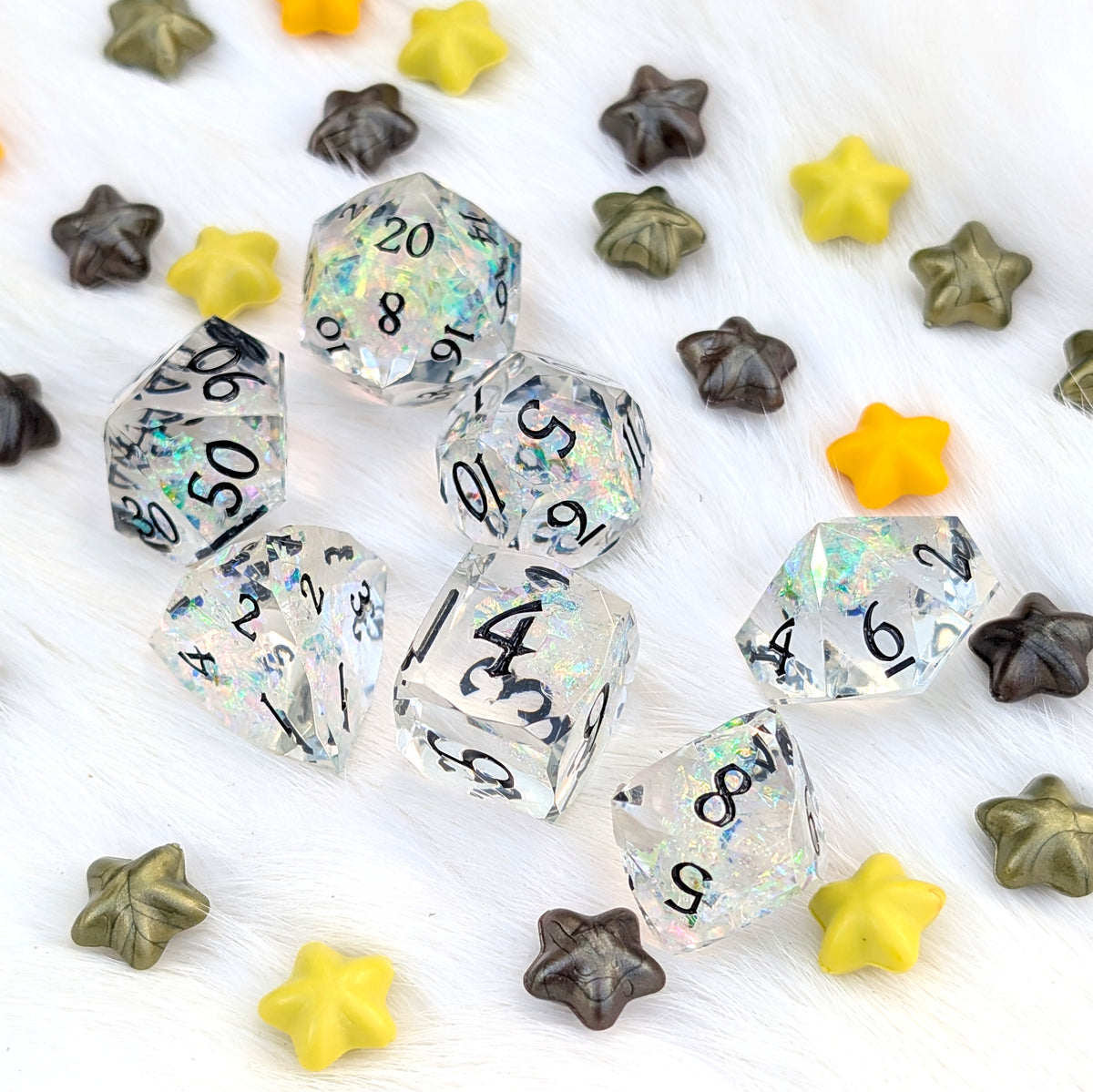 A set of clear acrylic polyhedral dice with black printed numbers and holographic iridescent flecks, scattered on a white faux fur background alongside yellow, brown, and olive star-shaped beads.