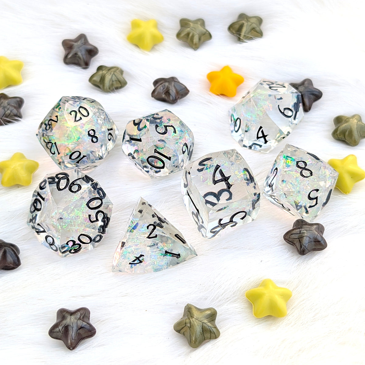 Clear acrylic polyhedral gaming dice with black printed numbers and iridescent glitter flecks, arranged on a white textured surface with small star-shaped beads in yellow, olive green, and dark brown.
