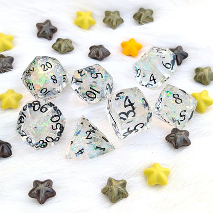 Clear acrylic polyhedral gaming dice with black printed numbers and iridescent glitter flecks, arranged on a white textured surface with small star-shaped beads in yellow, olive green, and dark brown.