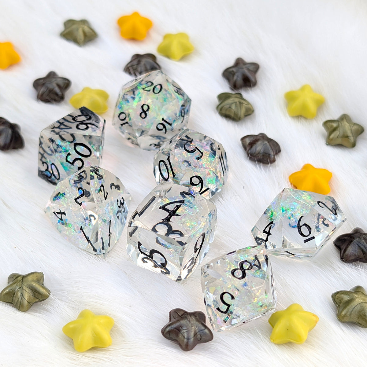 A set of clear acrylic polyhedral dice with black printed numbers and iridescent glitter, arranged on a white faux fur surface with scattered star-shaped decorative pieces in yellow, brown, and olive green.