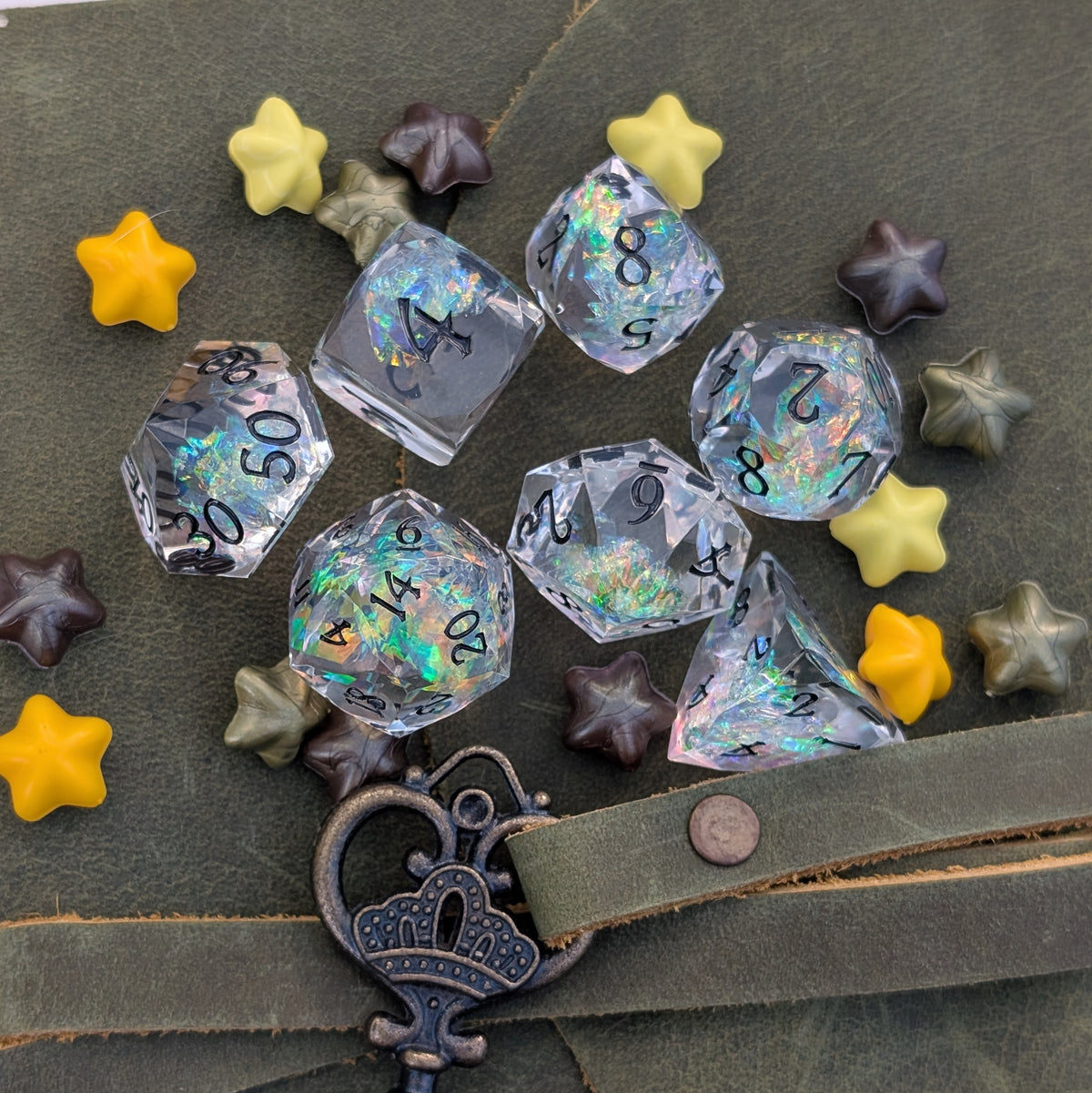 A set of translucent acrylic polyhedral dice with black printed numbers and iridescent holographic glitter, arranged on a dark olive green leather surface with decorative star-shaped beads and a metal key.