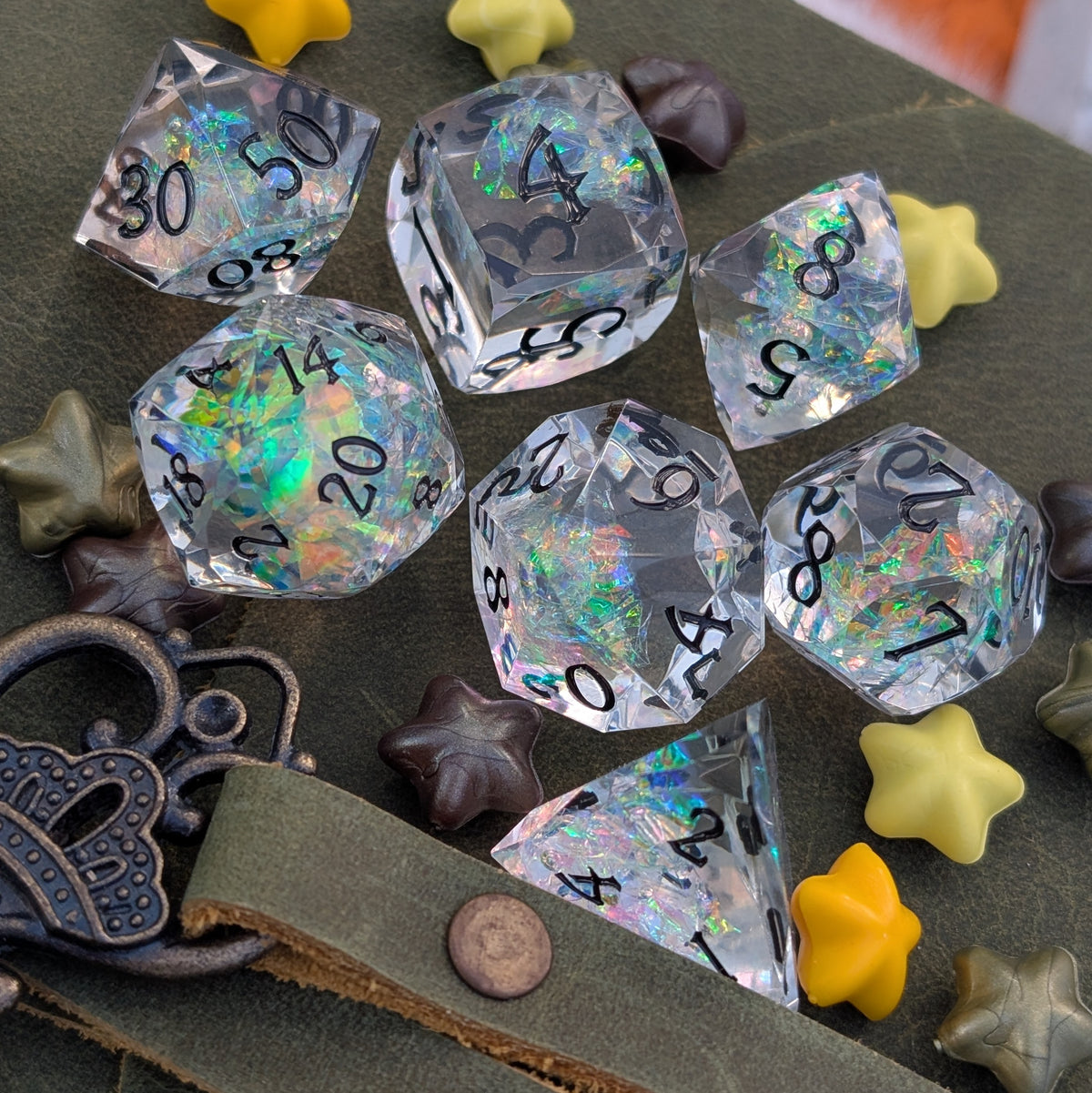 A set of clear acrylic polyhedral dice with iridescent opal-like glitter and black printed numbers, arranged on a dark olive surface with decorative star-shaped props and a metal ornament.