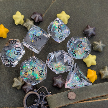 A set of clear acrylic polyhedral dice with black printed numbers and holographic iridescent glitter, arranged on a dark olive green textured surface with small star-shaped accessories.