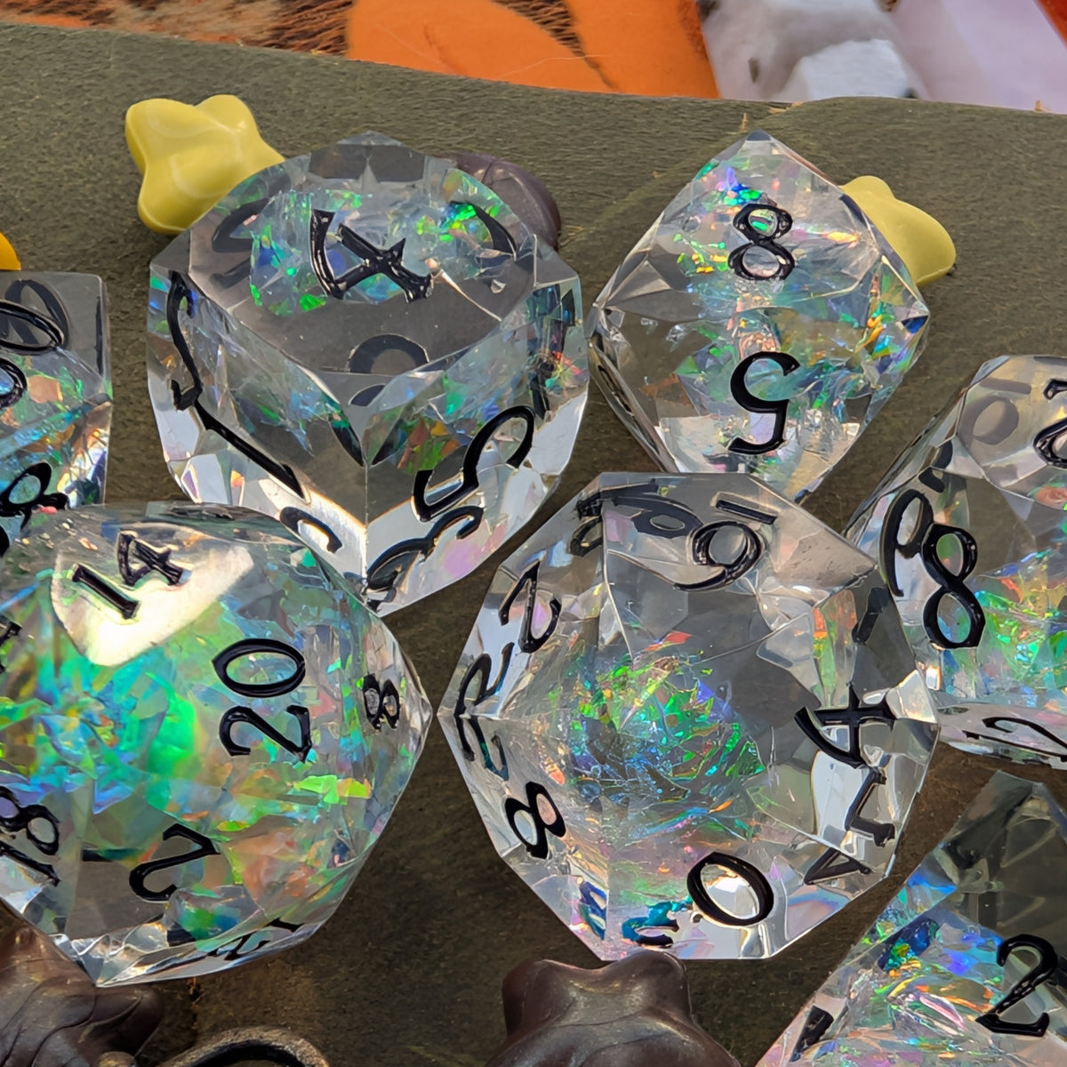 A set of transparent acrylic polyhedral dice with holographic iridescent glitter and black printed numbers, shown on a textured olive green surface.