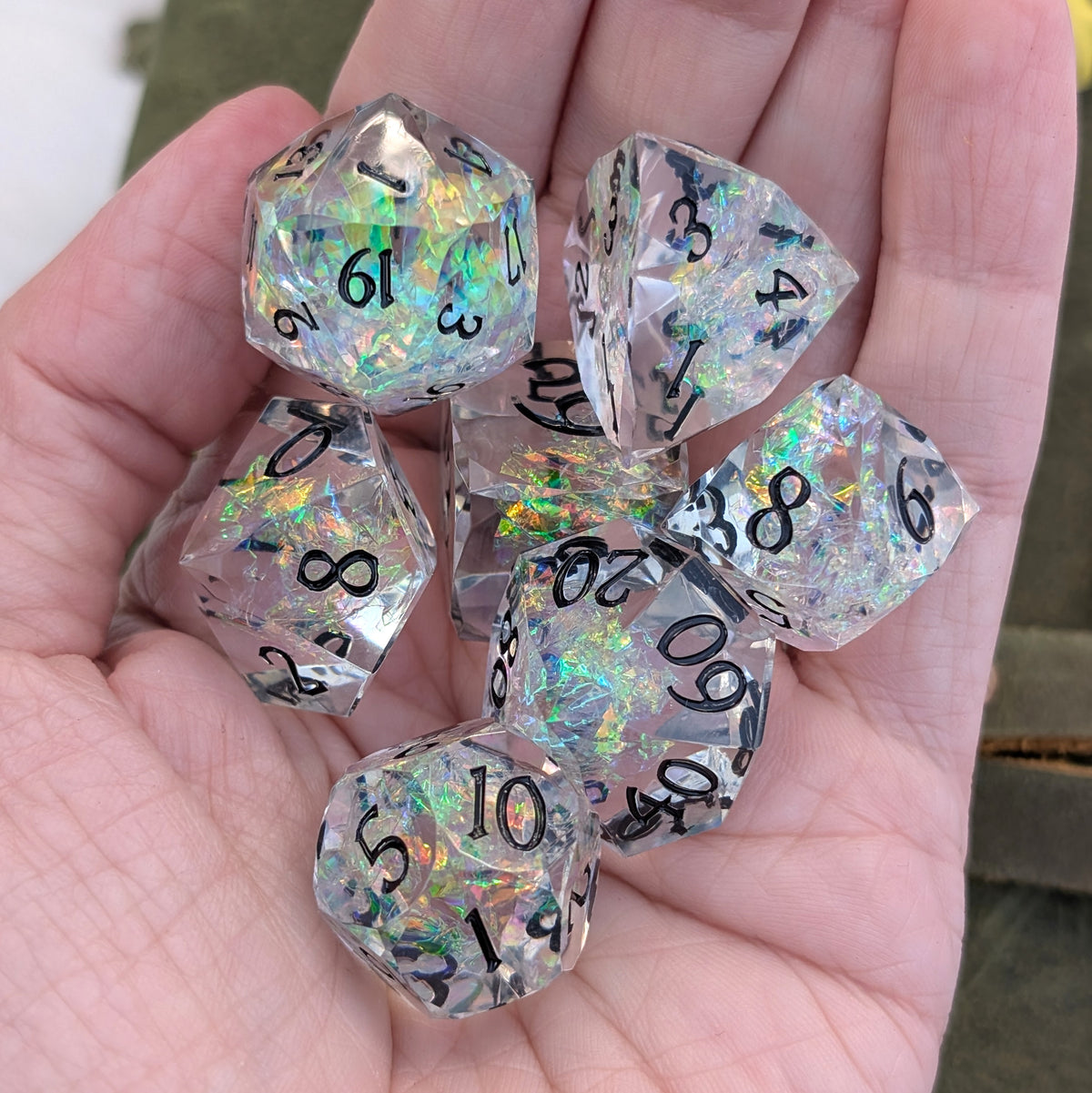 A set of clear holographic polyhedral dice with black printed numbers, held in a person's hand.