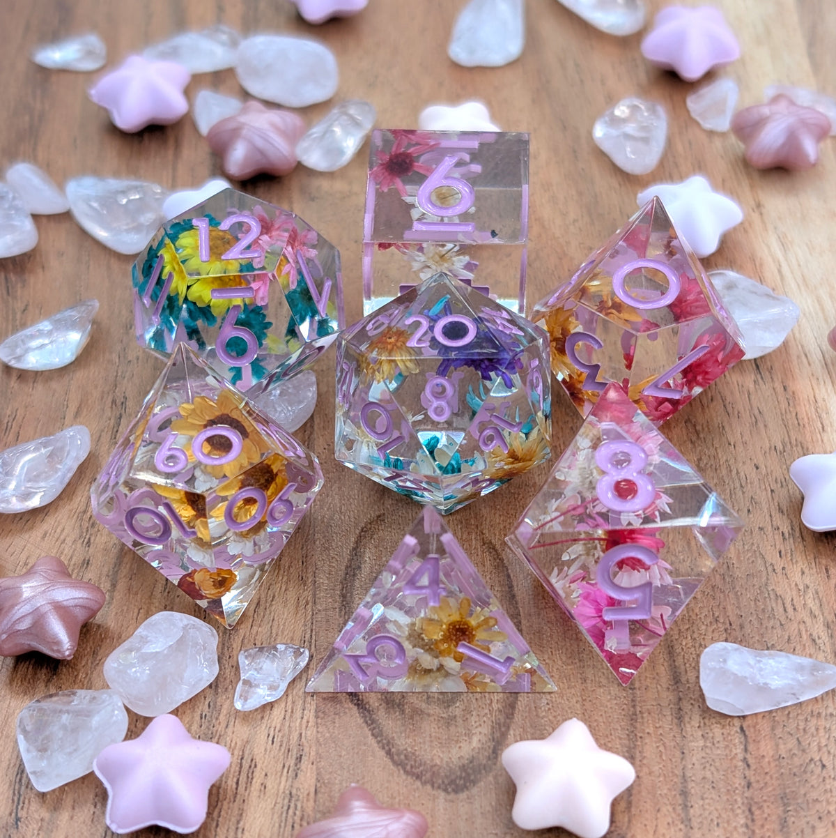 A set of translucent polyhedral dice with embedded flowers and glitter, displayed on a wooden surface surrounded by decorative pink and clear stones.