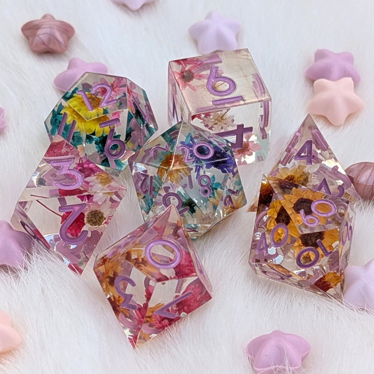 A set of translucent resin polyhedral dice with embedded dried flowers and purple numbers, arranged on a white faux fur surface with pastel star-shaped props.