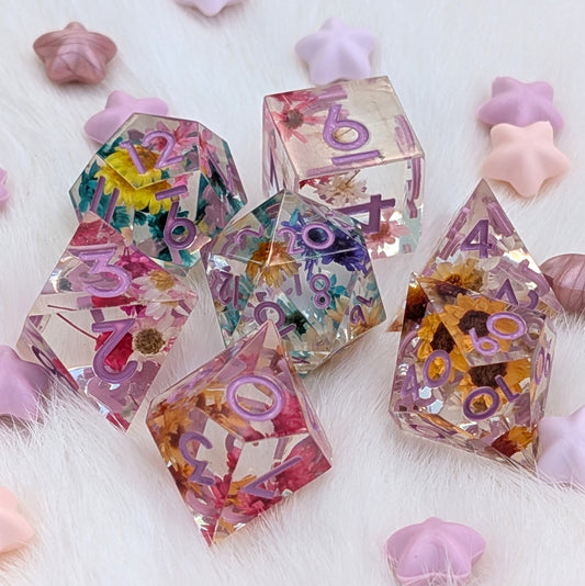A set of translucent resin polyhedral dice with embedded dried flowers and purple numbers, arranged on a white faux fur surface with pastel star-shaped props.