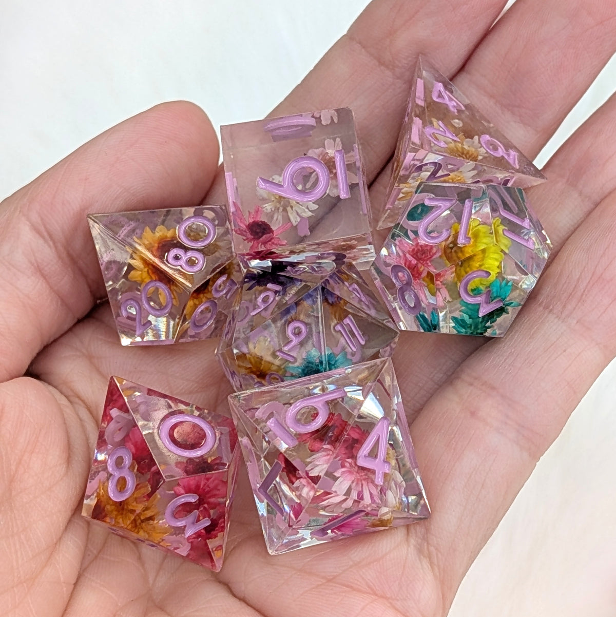 A set of translucent polyhedral dice with pink numbered faces and embedded dried flowers, held in a person's hand against a light background.