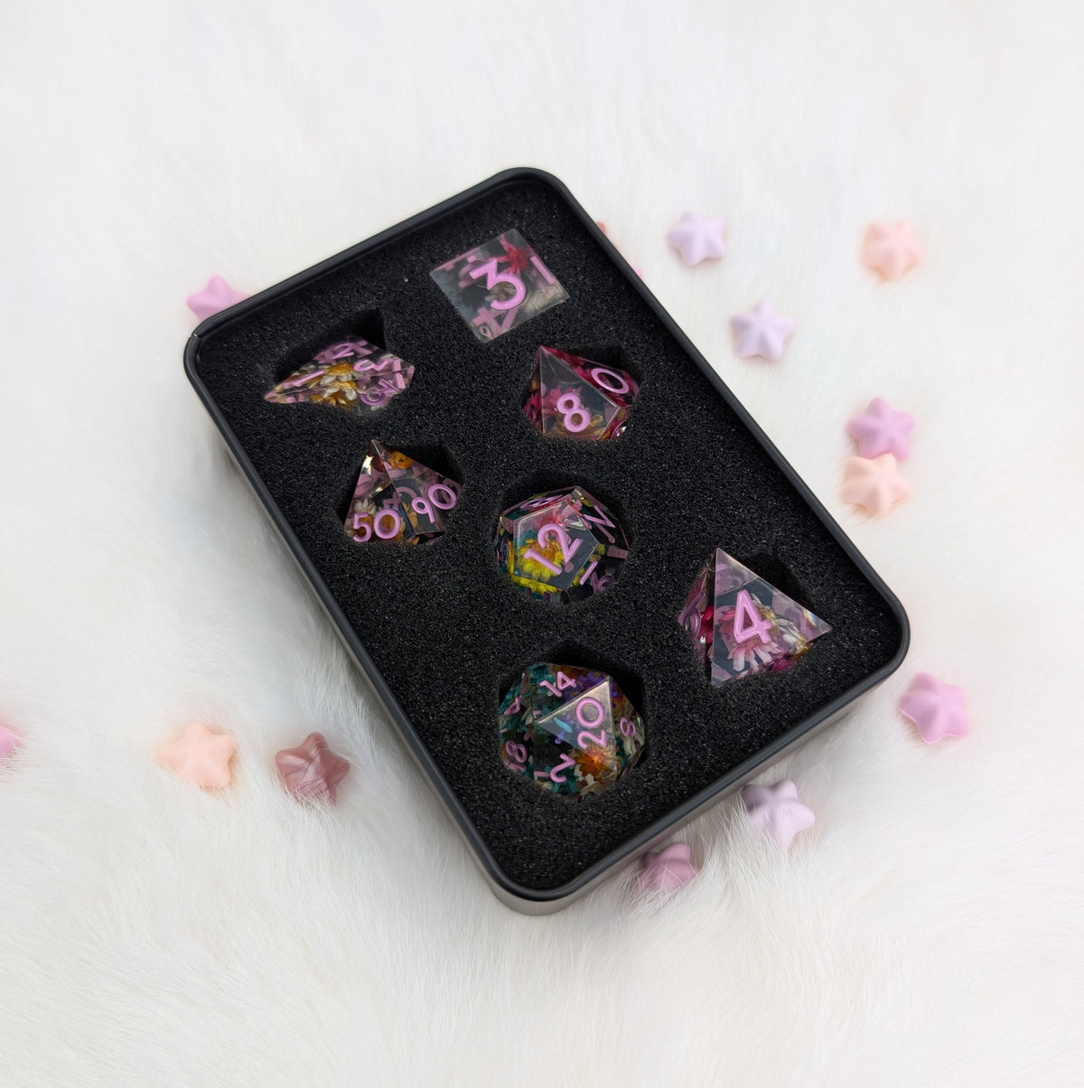 A set of multicolor polyhedral gaming dice displayed in a black foam tray, placed on a white faux fur surface with scattered pastel star-shaped decorations.
