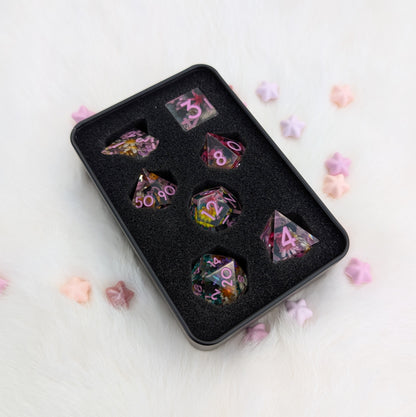 A set of multicolor polyhedral gaming dice displayed in a black foam tray, placed on a white faux fur surface with scattered pastel star-shaped decorations.