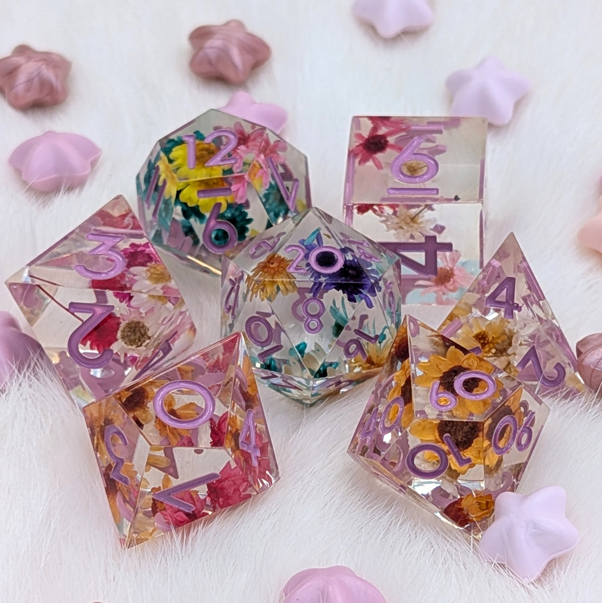 A set of transparent polyhedral dice with embedded dried flowers and purple printed numbers, arranged on a white faux-fur surface with pink star-shaped props.