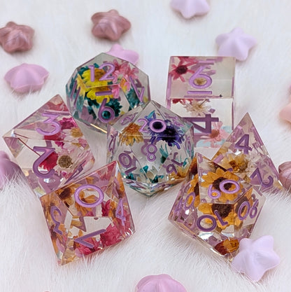 A set of transparent polyhedral dice with embedded dried flowers and purple printed numbers, arranged on a white faux-fur surface with pink star-shaped props.