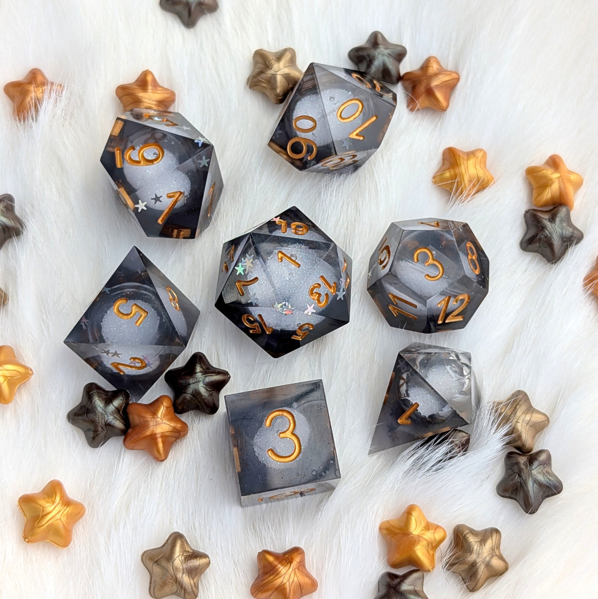 A set of translucent smoky gray polyhedral gaming dice with metallic gold numbers, arranged on a white faux fur surface with scattered star-shaped decorative pieces in gold, bronze, and dark gray.