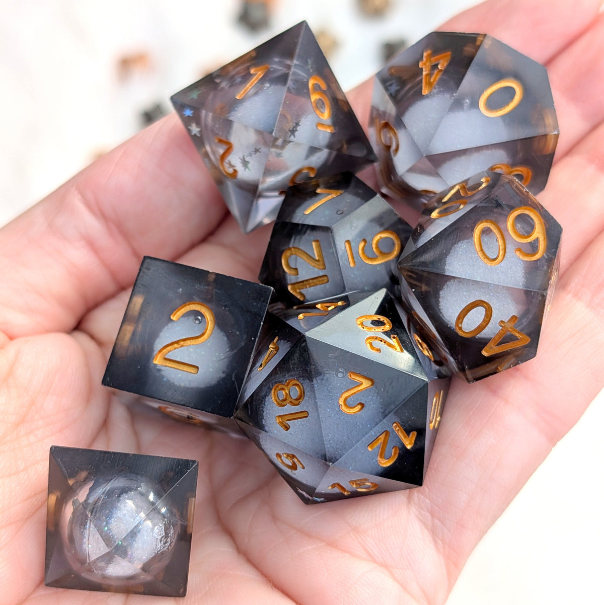 A set of translucent smoky gray polyhedral gaming dice with metallic gold numbers, shown resting in a person's hand. A single die is placed in front on a white surface.