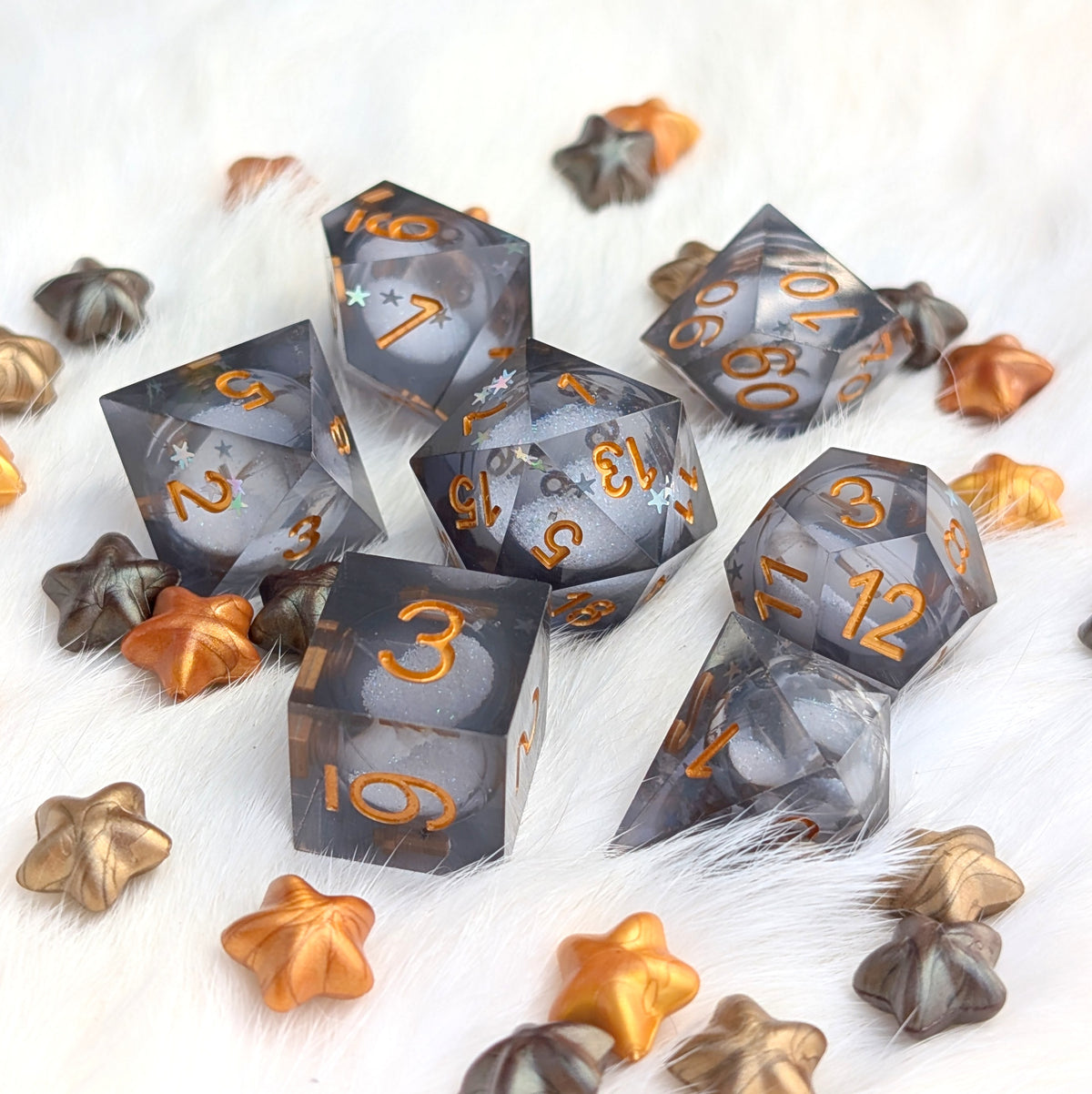 A set of translucent smoky gray polyhedral gaming dice with metallic gold numbers, arranged on a white faux fur surface with decorative star-shaped ornaments.