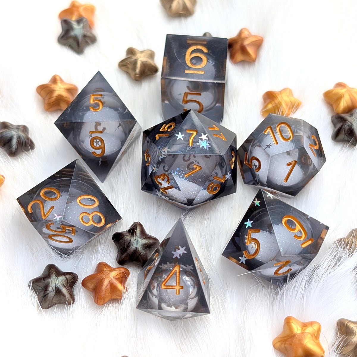 A set of translucent smoky gray polyhedral dice with metallic gold numbers, arranged on a white faux-fur surface with scattered star-shaped decorative pieces in gold, bronze, and dark gray.