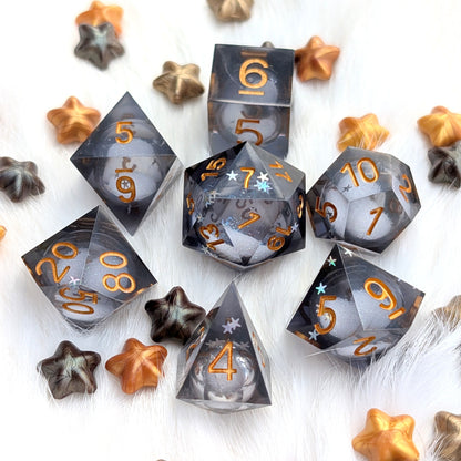 A set of translucent smoky gray polyhedral dice with metallic gold numbers, arranged on a white faux-fur surface with scattered star-shaped decorative pieces in gold, bronze, and dark gray.