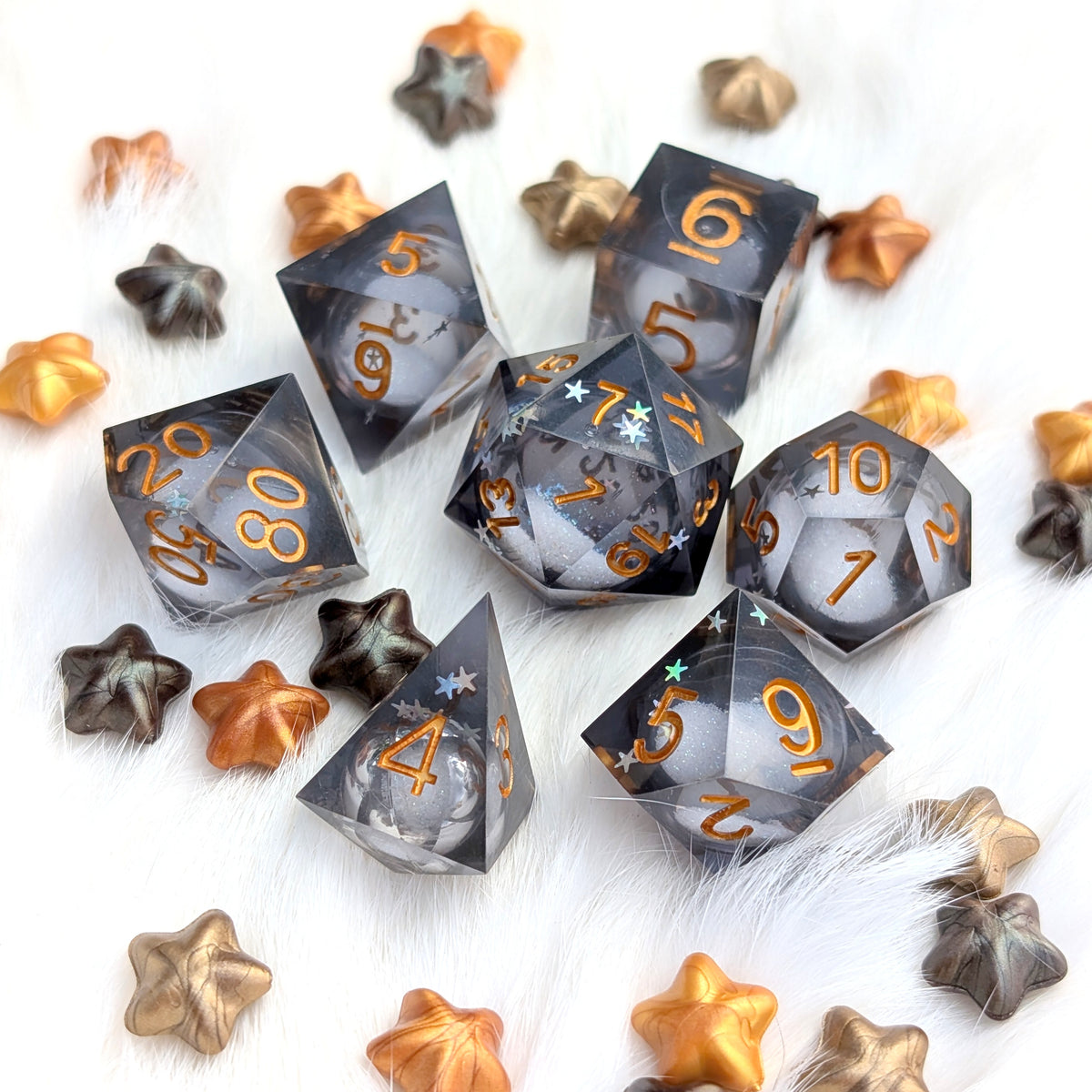 A set of translucent smoky gray polyhedral gaming dice with metallic gold numbers, arranged on a white faux fur surface with scattered metallic gold and bronze star-shaped props.
