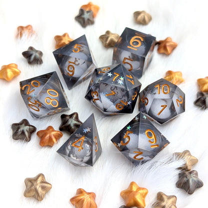 A set of translucent smoky gray polyhedral gaming dice with metallic gold numbers, arranged on a white faux fur surface with scattered metallic gold and bronze star-shaped props.
