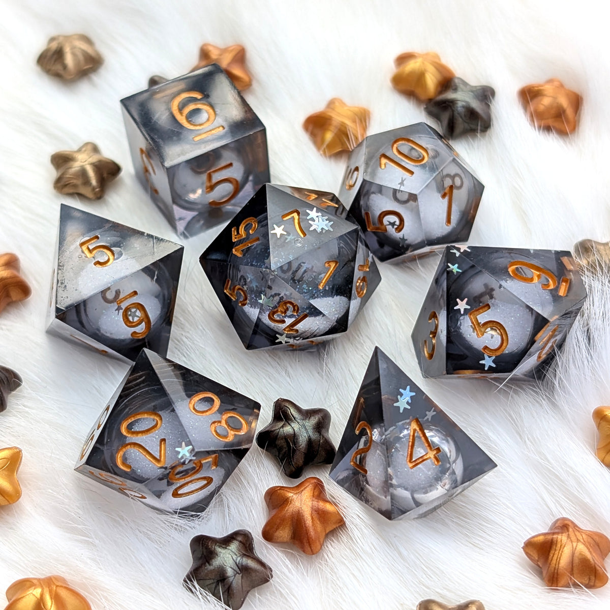 A set of translucent smoky gray polyhedral gaming dice with metallic gold numbers, arranged on a white faux fur surface with scattered bronze and dark metallic star-shaped decorative pieces.