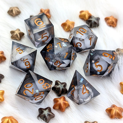 A set of translucent smoky gray polyhedral gaming dice with metallic gold numbers, arranged on a white faux fur surface with scattered bronze and dark metallic star-shaped decorative pieces.