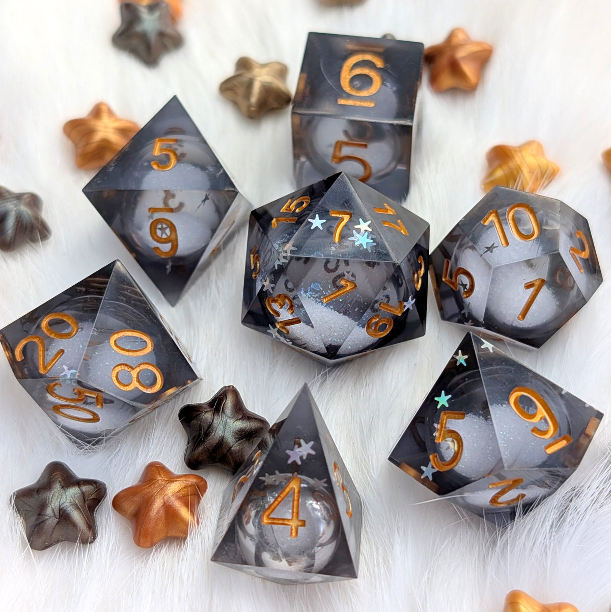 A set of translucent smoky gray polyhedral gaming dice with metallic gold numbers, arranged on a white faux fur surface with small metallic star-shaped props.