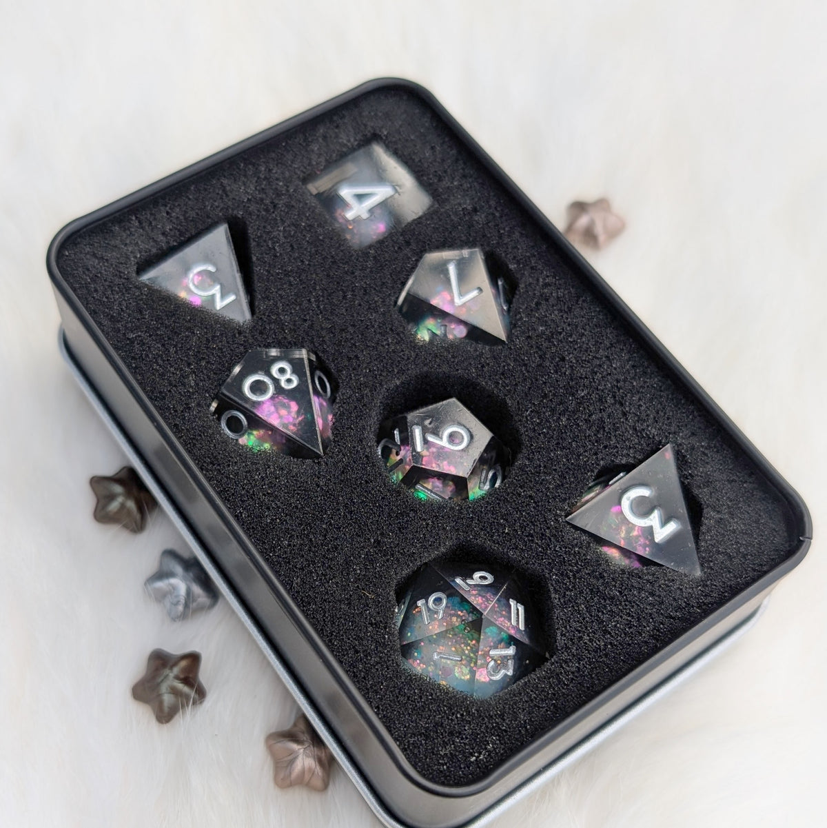 A set of seven transparent acrylic polyhedral dice with colorful glitter and floral patterns, displayed inside a black metal tin with a black foam insert. The tin is placed on a white textured surface with a few decorative star-shaped items nearby.