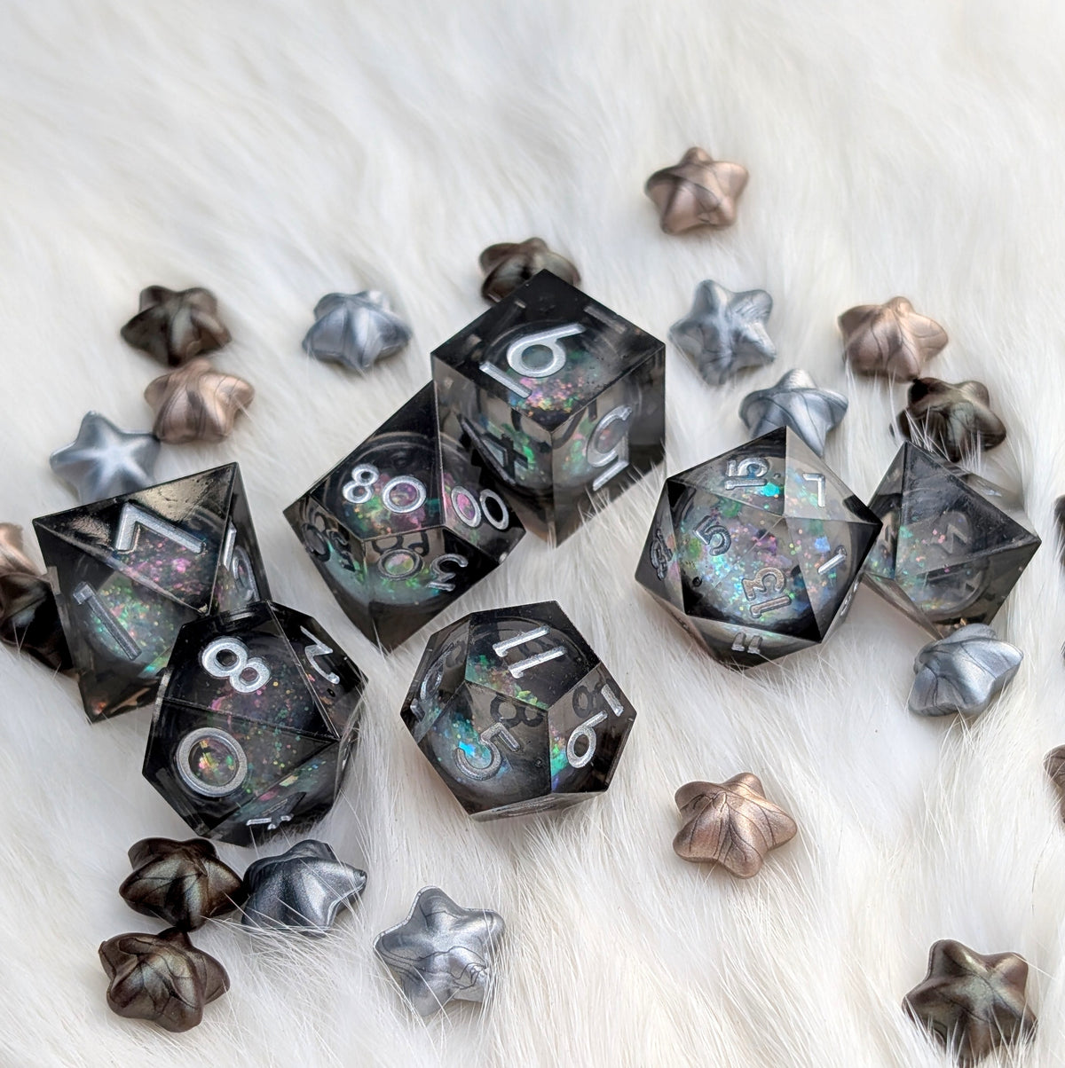 A set of transparent black polyhedral gaming dice with iridescent glitter and white numbering, arranged on a white faux fur surface with matching silver and bronze star-shaped accessories.