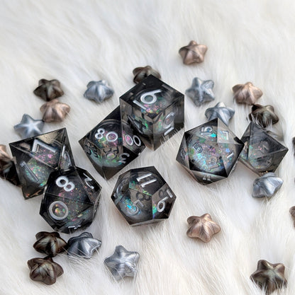 A set of transparent black polyhedral gaming dice with iridescent glitter and white numbering, arranged on a white faux fur surface with matching silver and bronze star-shaped accessories.