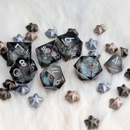 A set of translucent acrylic polyhedral dice with iridescent glitter and star-shaped dice in metallic silver, bronze, and rose gold, arranged on a white faux fur background.