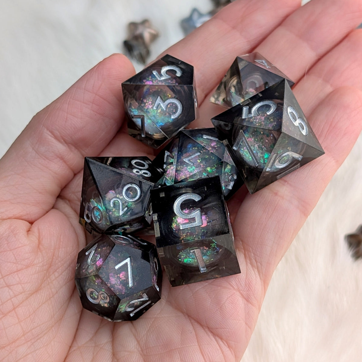 A set of seven black polyhedral gaming dice with holographic glitter and white numbering, held in a person's hand against a soft white background.