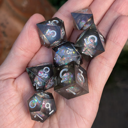 A set of seven translucent dark gray polyhedral gaming dice with holographic glitter and white numbering, held in a person's hand.