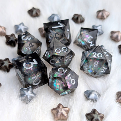 A set of translucent black polyhedral gaming dice with holographic glitter and silver numbering, arranged on a white faux fur surface with scattered metallic star-shaped props.