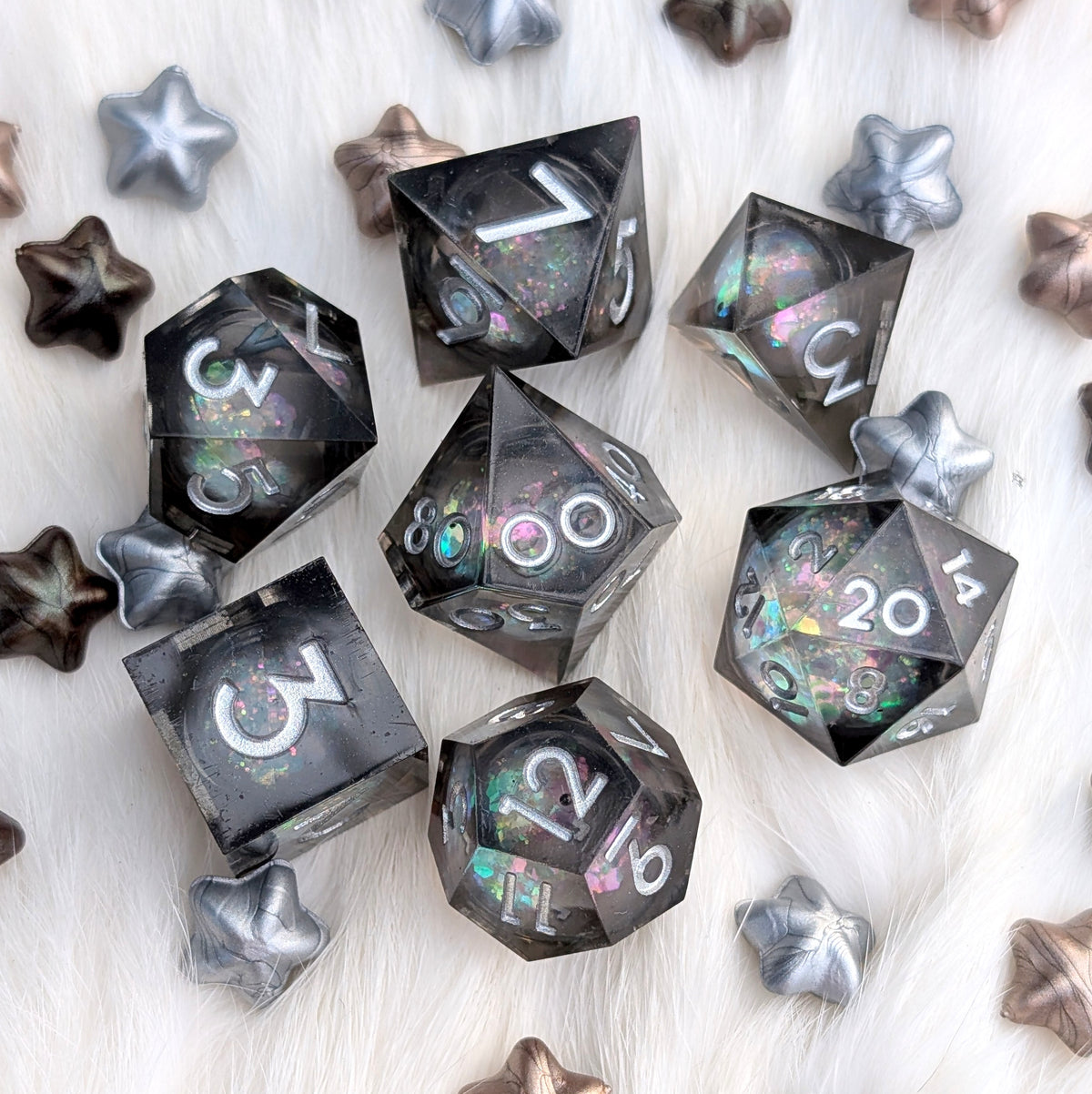 A set of translucent smoky gray polyhedral gaming dice with holographic glitter and white numbering, arranged on a white faux fur surface with scattered metallic star-shaped props.
